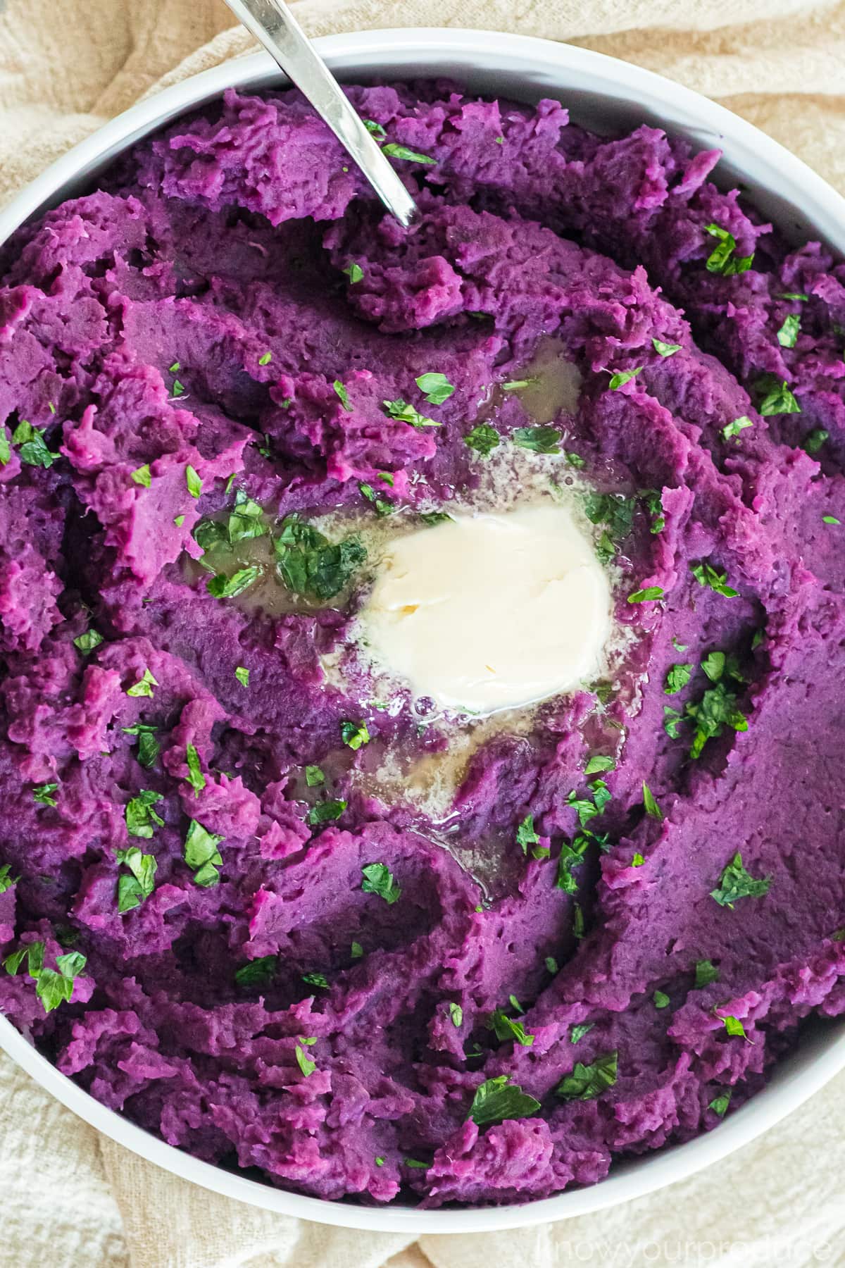 close up of mashed purple sweet potatoes with a pat of butter and parsley garnish in a white bowl with a spoon on the top