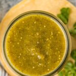 overhead photo of green enchilada sauce in a glass mason jar with cilantro, lime, and garlic scattered around.