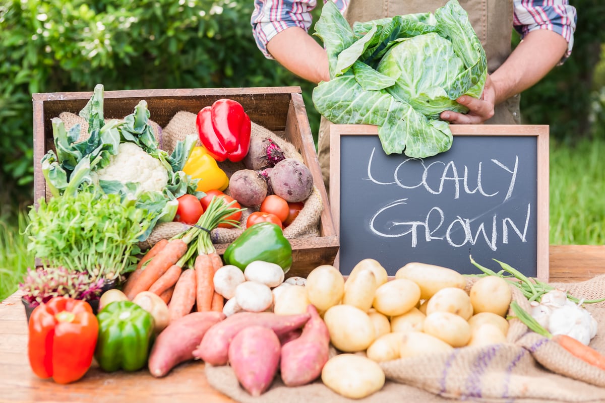 farmer holding cabbage head with locally grown produce sign and produce on a table.