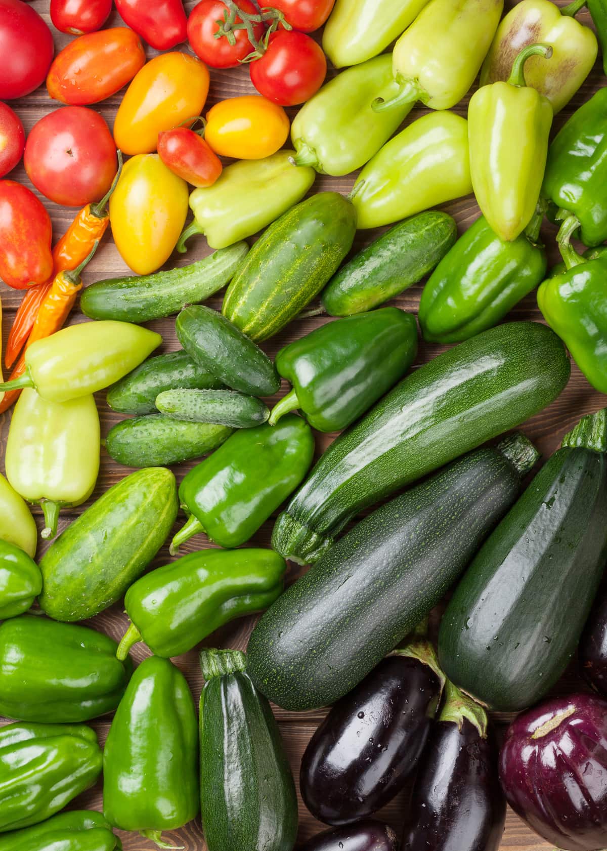 fresh tomatoes, peppers, cucumbers, zucchini, and eggplant  on a table in color gradient from red, yellow, green, then purple.