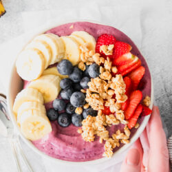 hand holding a white bowl that is filled with a thick acai smoothie topped with sliced banana, granola, blueberries, and sliced strawberries.