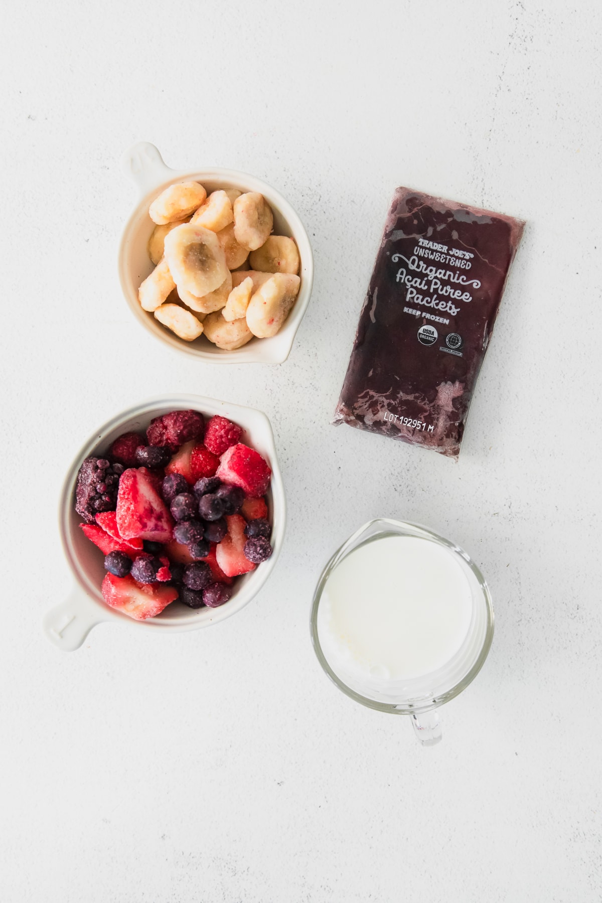 overhead photo of acai bowl ingredients; acai puree packet, frozen berries in a white bowl, frozen banana sliced in a white bowl, and a measuring cup filled with oat milk.