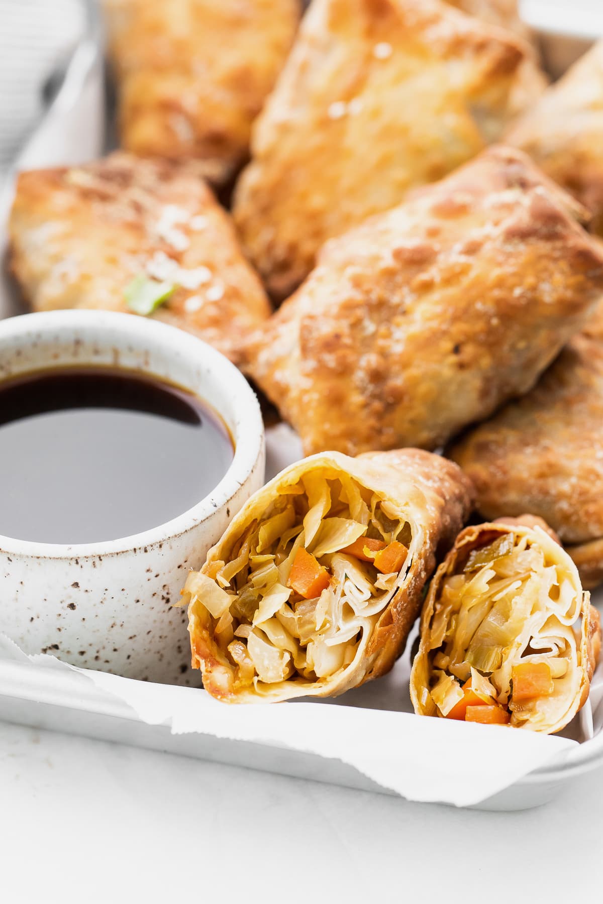 air fryer egg roll sliced in half close up with sauce in a white bowl next to it and more egg rolls behind it on a tray.