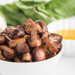 white bowl with cooked balsamic mushrooms and a bowl of oil in the background as well as herbs.