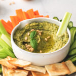 pesto hummus in a white low bowl with sliced cucumbers, celery, carrots, and pita chips around it.