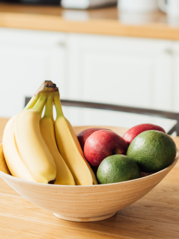 wooden basket with bananas, apples, and avocado on a countertop showing how to store avocado and other fruits to ripen the avocados.