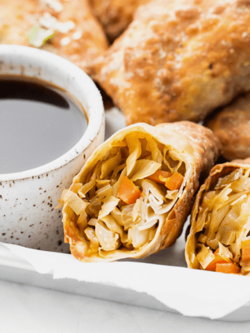 air fryer egg roll sliced in half close up with sauce in a white bowl next to it and more egg rolls behind it on a tray.