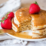 dairy free pancakes covered in syrup on a white plate and a strawberry on top of the stack of pancakes.