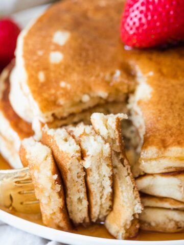 dairy free pancakes covered in syrup on a white plate with a fork holding a bite and a strawberry on top of the stack of pancakes.