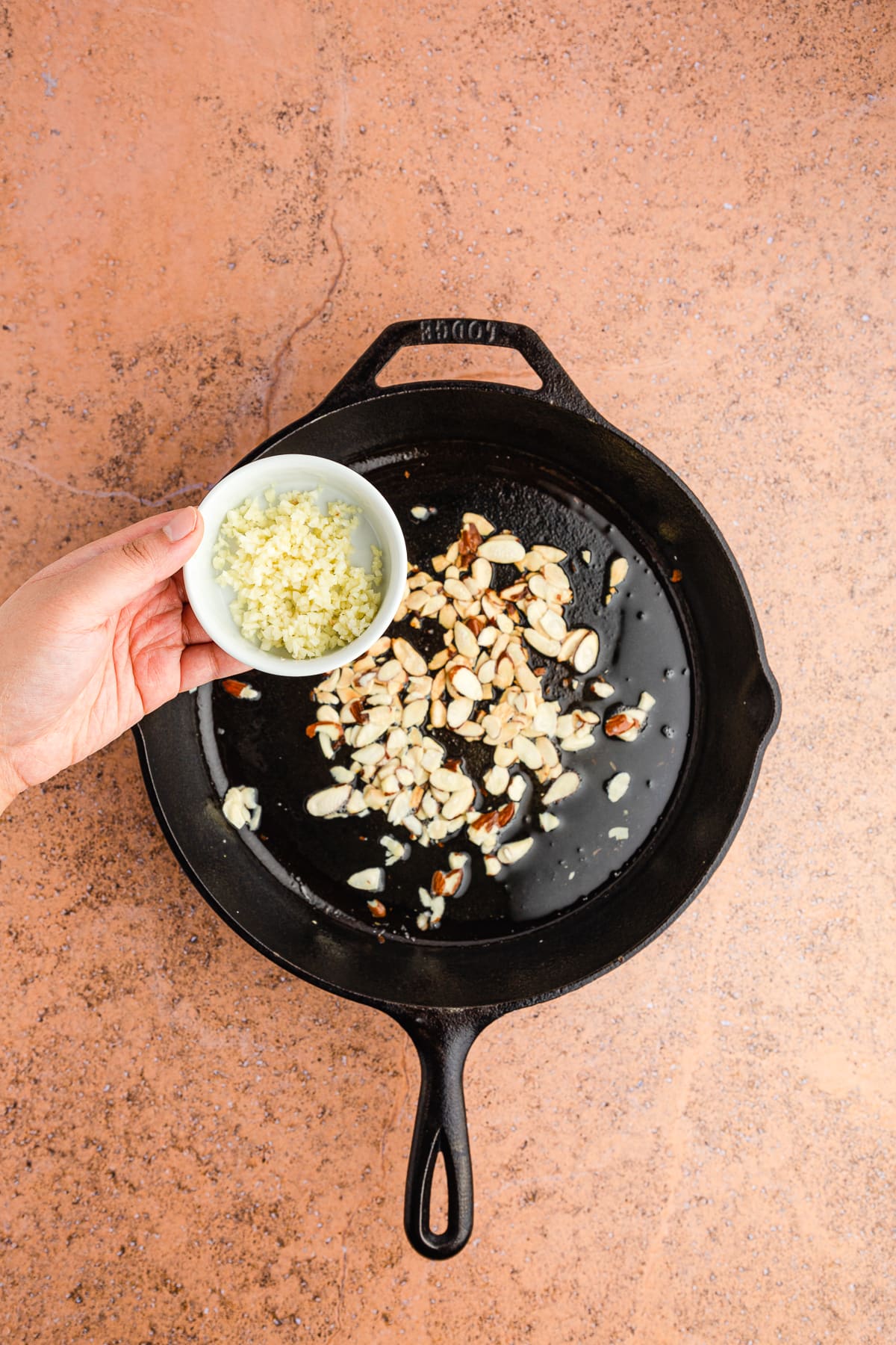 hand holding minced garlic in a bowl over a cast iron skillet with toasting slivered almonds.