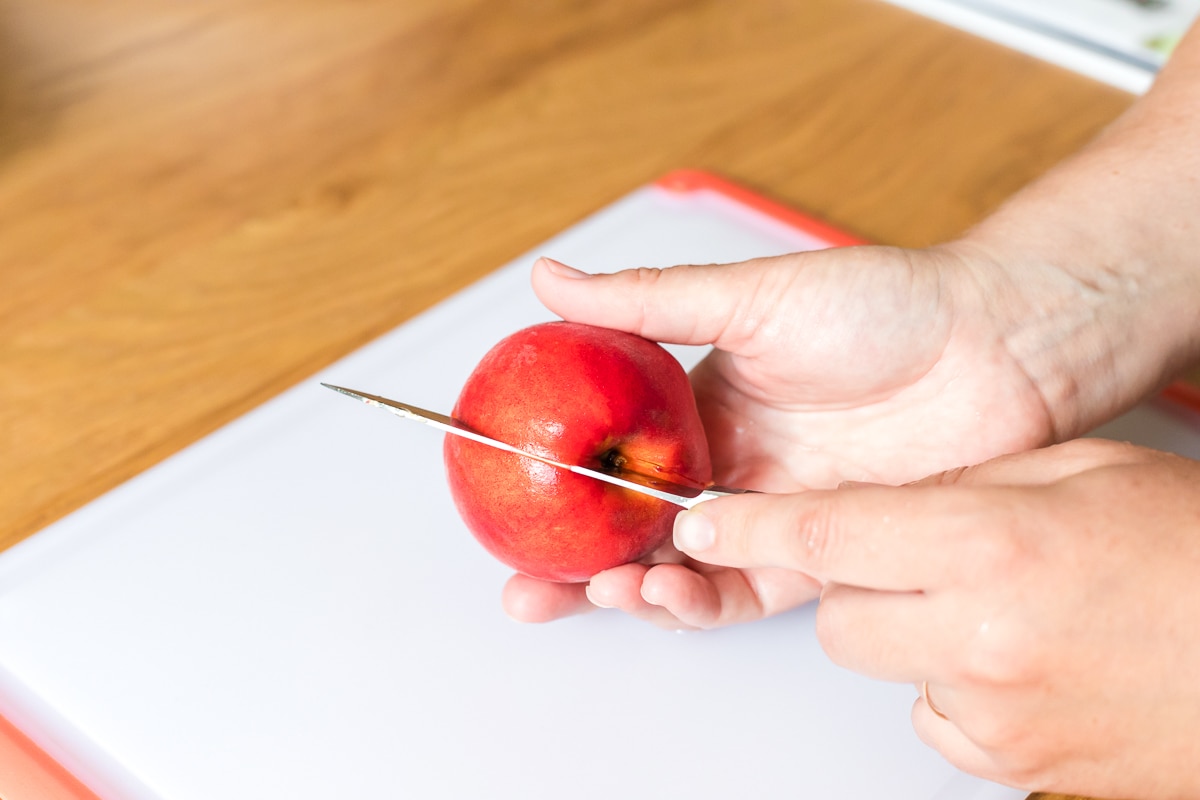 hand holding a peach and a paring knife scoring into the center of the peach.