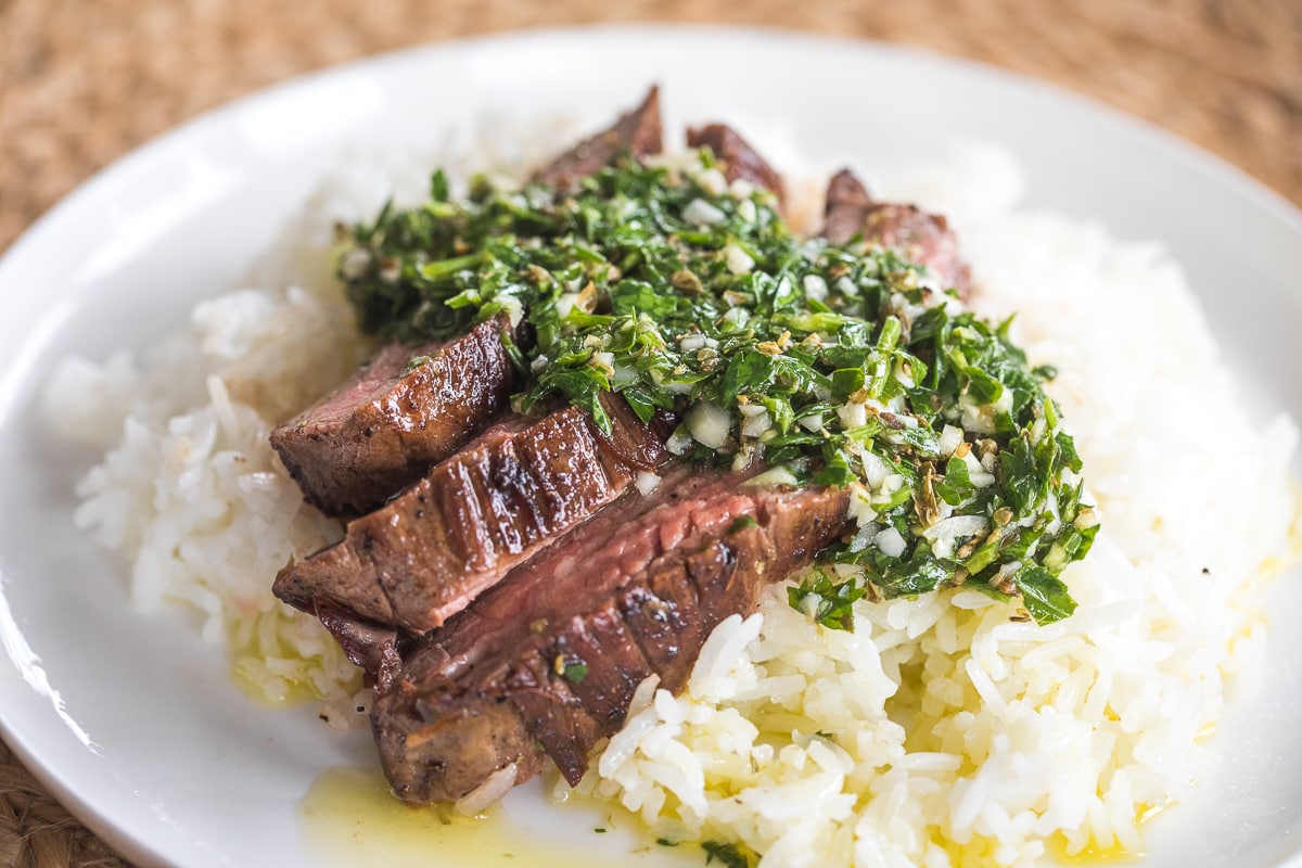 sliced flank steak with chimichurri sauce on top over rice on a white plate.