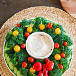 Christmas veggie tray on a white plate consistening of cucumber, broccoli, cherry tomatoes, with sour cream, a red pepper bow and onion dip.