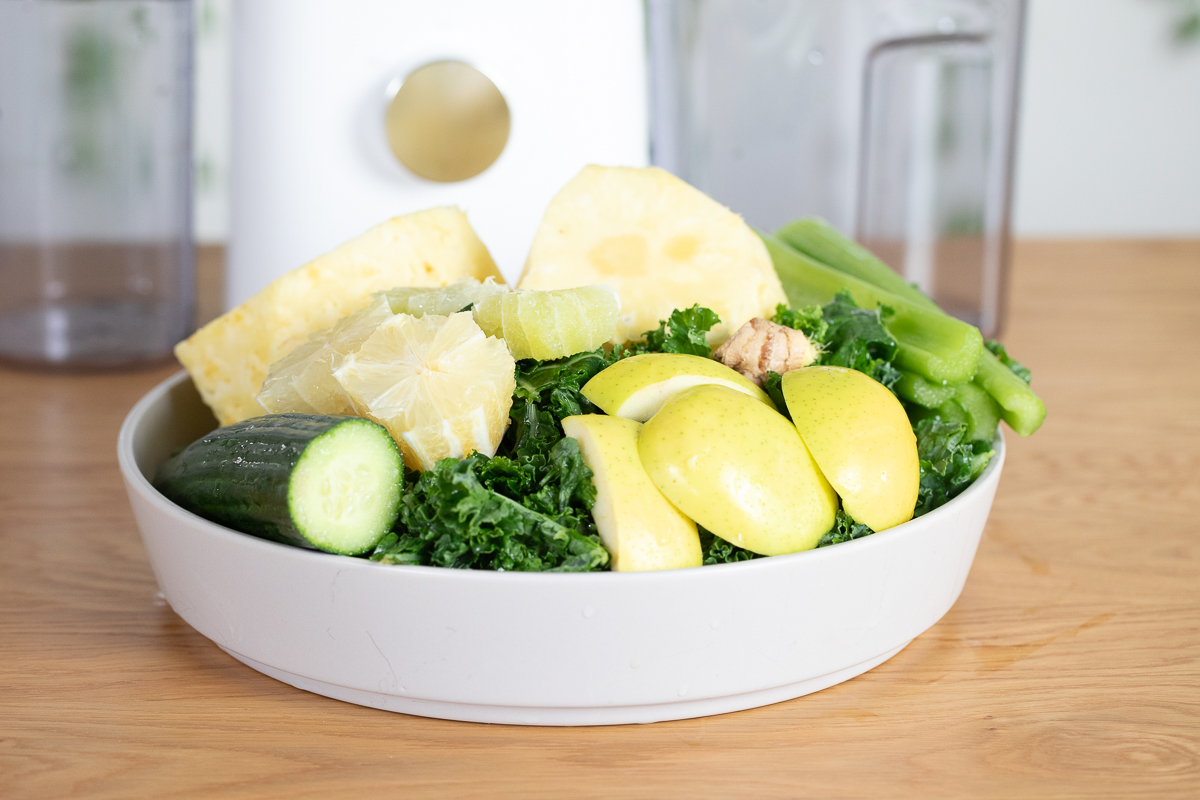 vertical photo of fresh vegetables and fruit prepped and ready for juicing in a serving bowl with a juicer behind it.