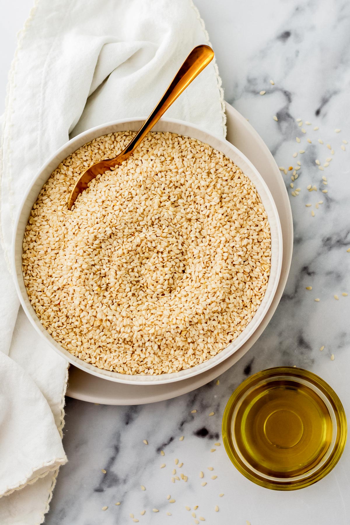 sesame seeds in a white bowl with a gold spoon and a little glass bowl below it with oil.