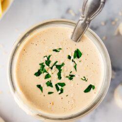 overhead photo of tahini sauce in a glass jar with a spoon in it and garnished on top with chopped parsley and sesame seeds.