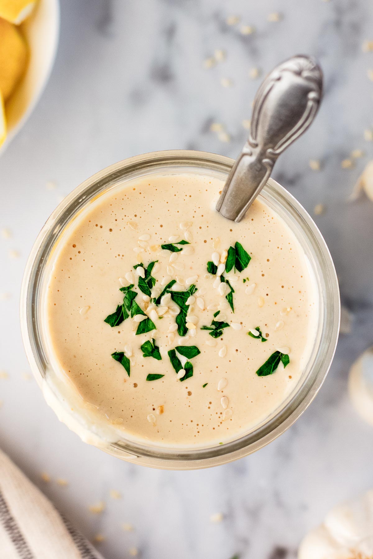 overhead photo of tahini sauce in a glass jar with a spoon in it and garnished on top with chopped parsley and sesame seeds.
