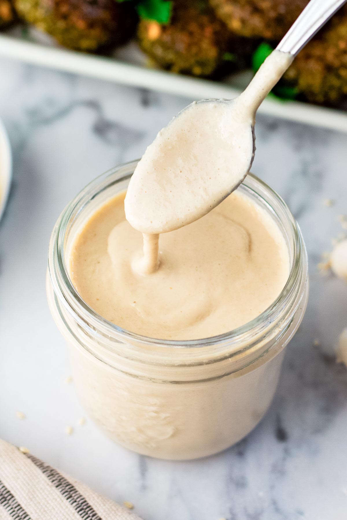 tahini sauce in a glass mason jar with a spoon in it drizzling the tahini sauce back in.