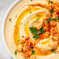 close up photo of hummus in a white low bowl garnished with spices, chickpeas, and freshly chopped parsley.