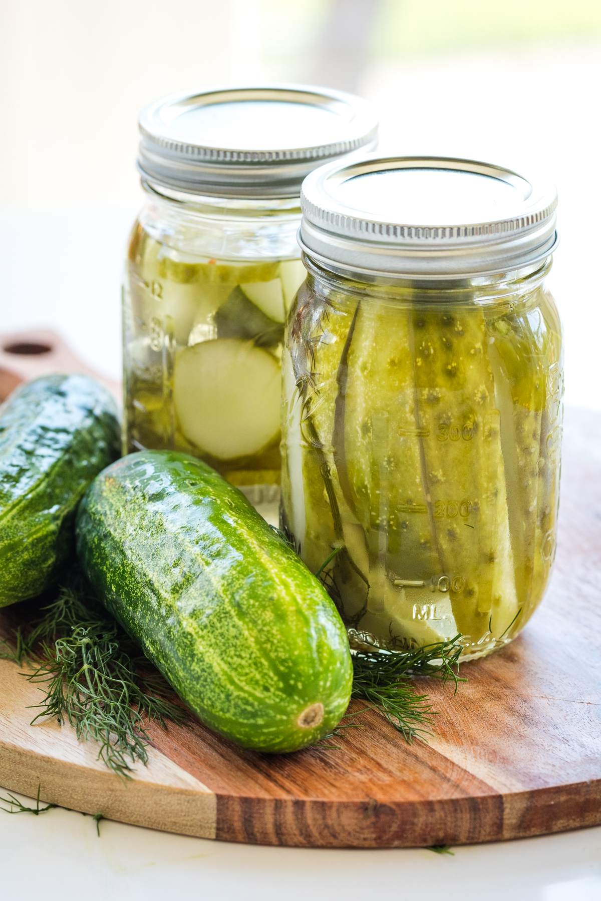 refrigerator dill pickles in mason jars with fresh kirby cucumbers and dill to the left on a wooden board.