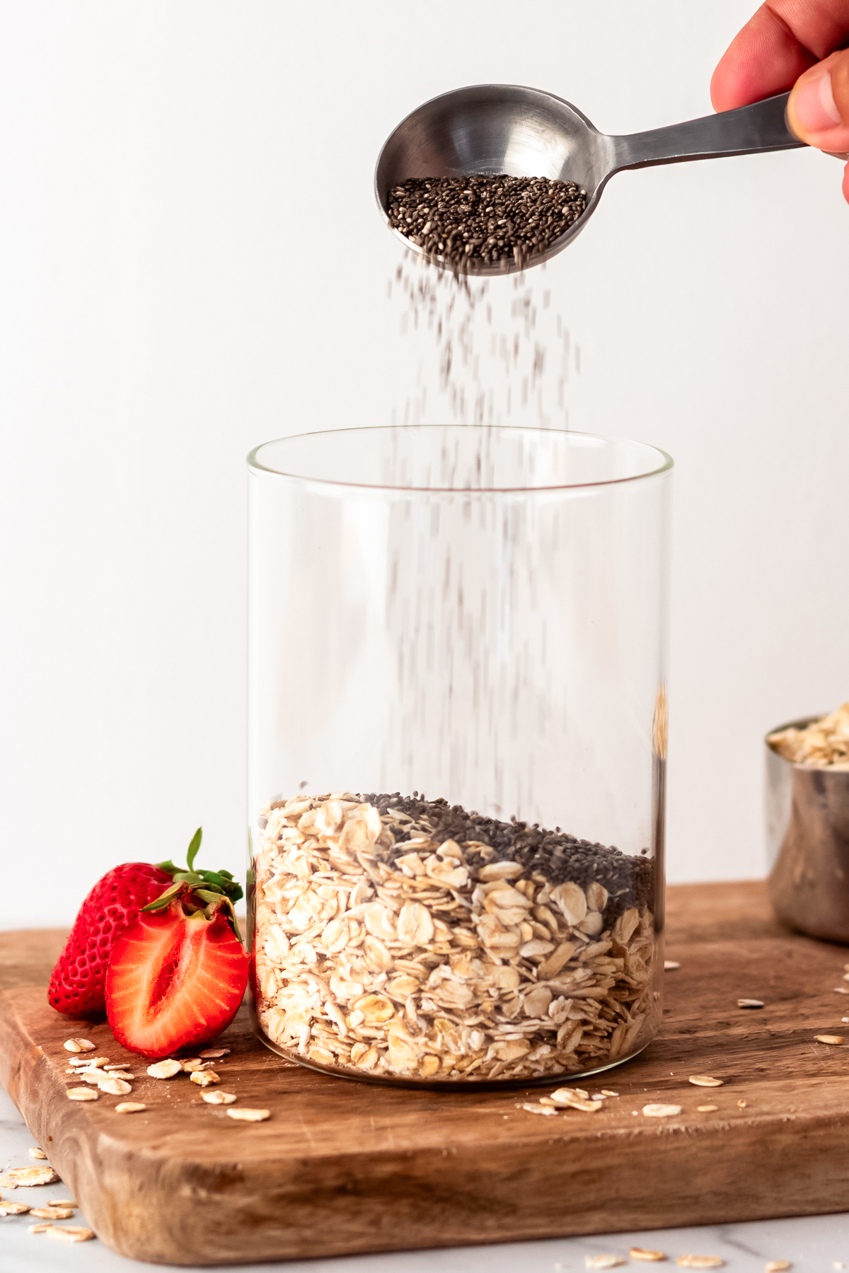 hand pouring chia seeds from measuring spoon into glass jar with oats in it.
