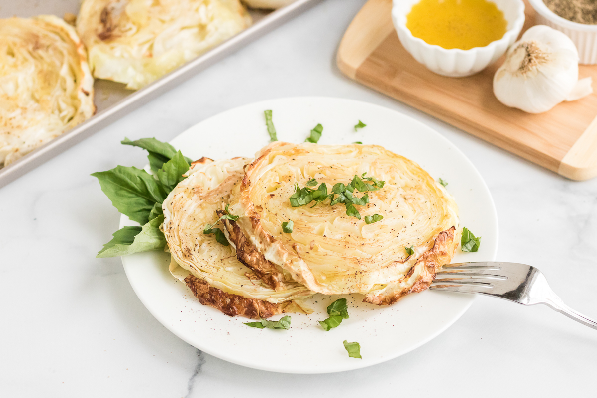 Roasted cabbage steaks served on a white plate with basil leaves and a fork, tray of cabbage steaks in the background.
