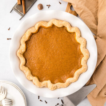 Overhead view of a whole pumpkin pie in a white dish surrounded by forks and fall spices.
