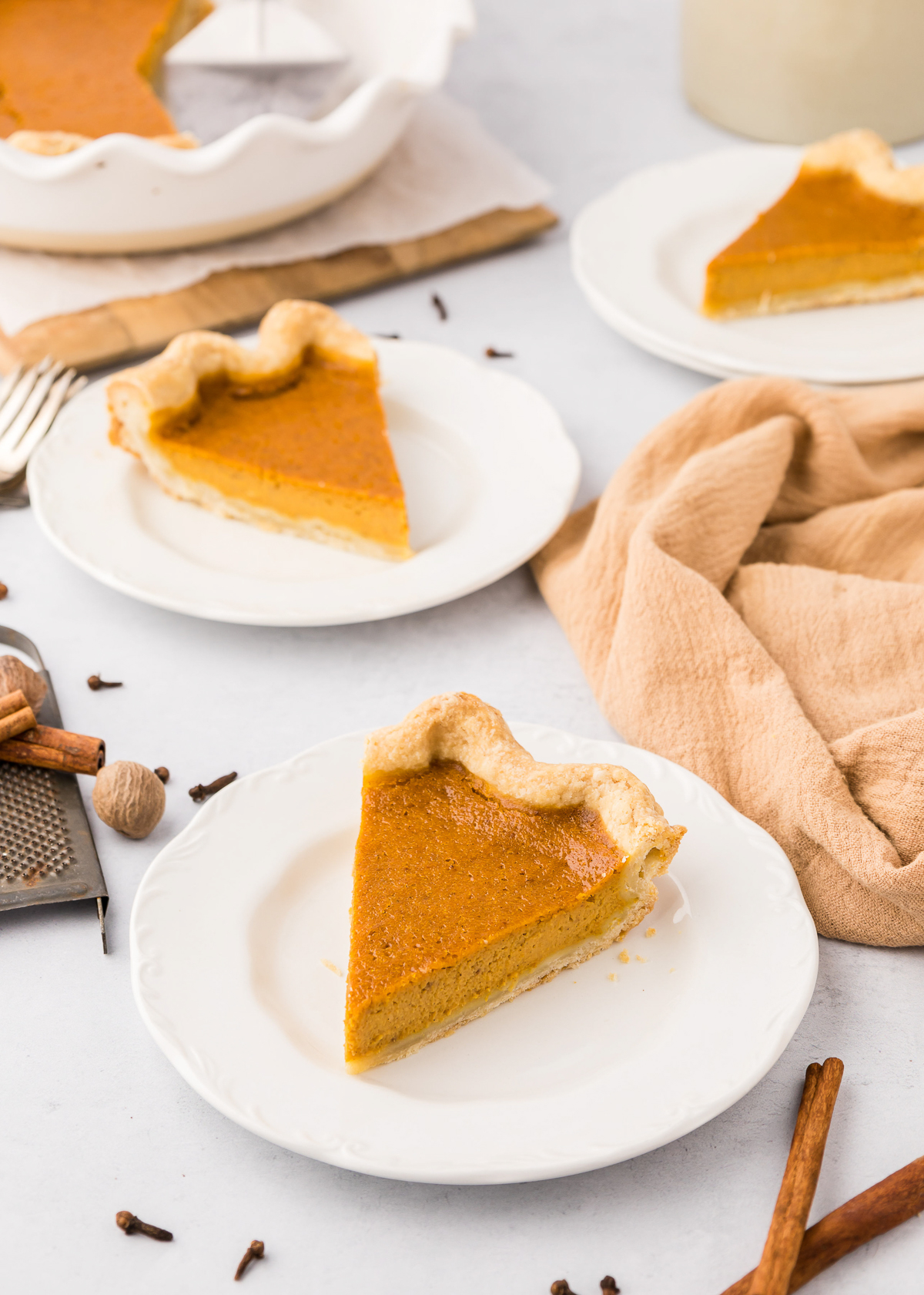 Two slices of pumpkin pie on white plates with spices and a fall linen cloth nearby.